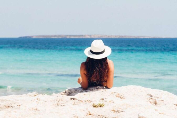 Une femme portant un chapeau blanc est assise sur une roche face à la mer, illustrant les dangers naturels à la plage.